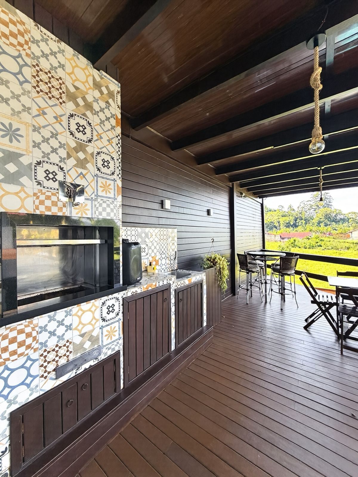 Churrasqueira da área de festas revestida de azulejos hidráulicos, com balcão, pia e deck de madeira com vista para o verde