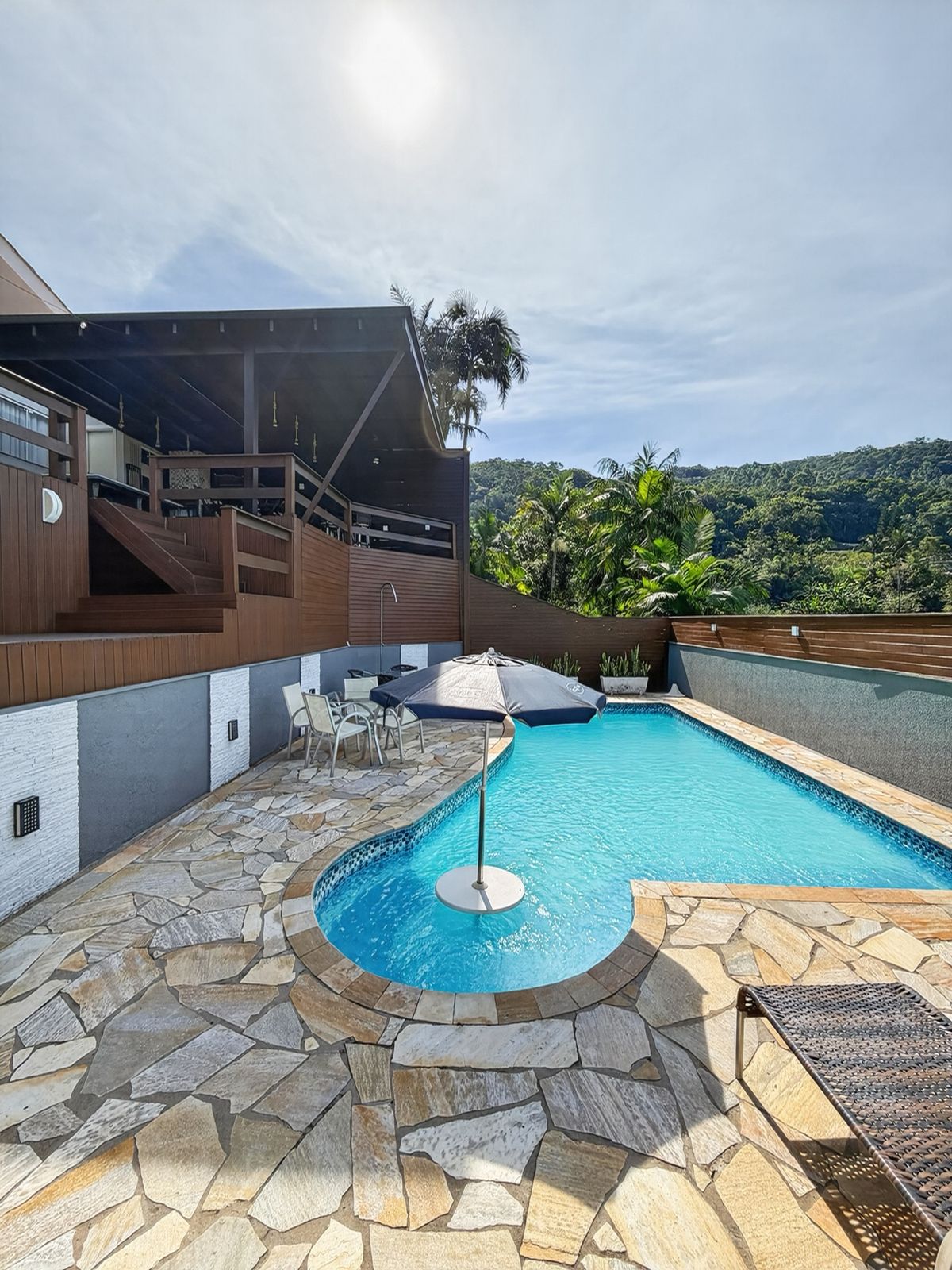 Piscina com ombrelone e mesas na área de pedra, abaixo do deck da área de festas