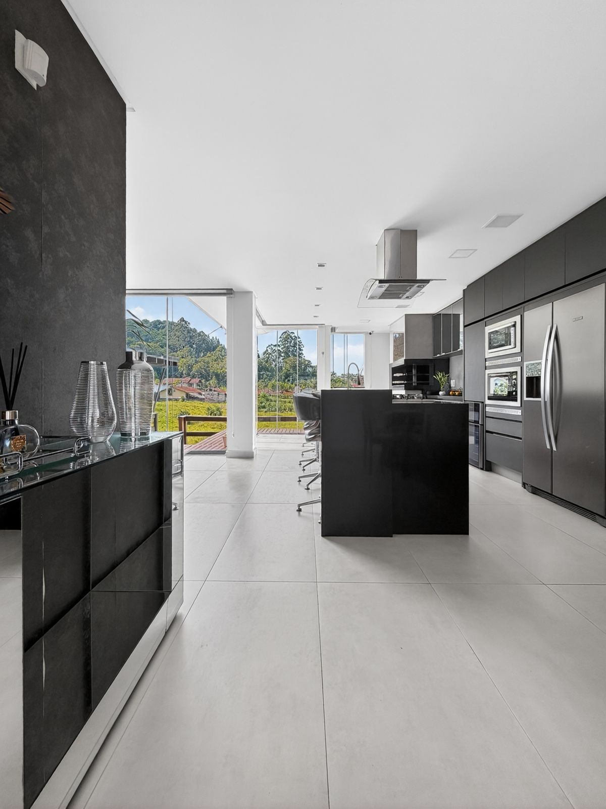 Cozinha gourmet integrada com ilha preta, torre de eletros inox e janelas de vidro com vista para o vale